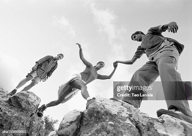 three young people climbing on rocks, woman being aided, low angle view, b&w. - black and white people holding hands stock pictures, royalty-free photos & images