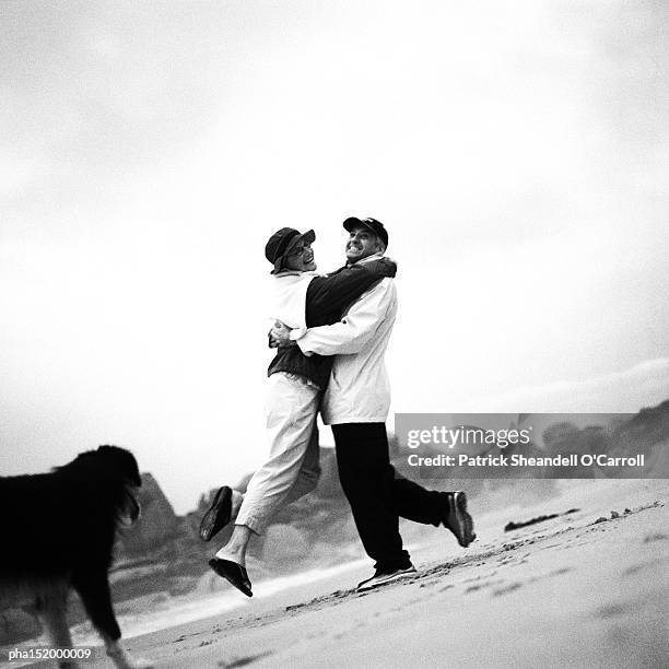 mature couple and dog on beach, woman jumping into man's arms, b&w. - joined at hip stock pictures, royalty-free photos & images