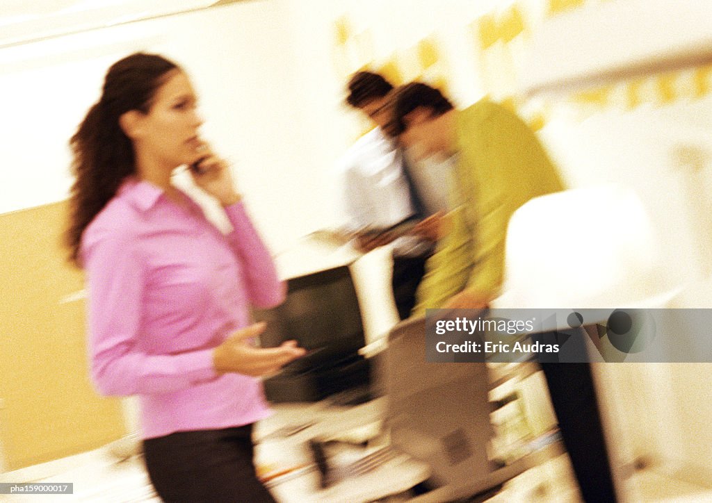 Woman standing, using cell phone, colleagues in background, blurred, tilt