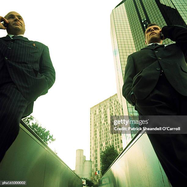 two businessmen on cell phones standing with hands in pocket, buildings in background, low angle view. - o foto e immagini stock