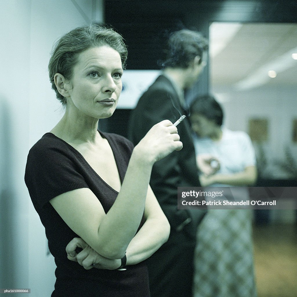 Woman smoking, arms crossed, people in background.