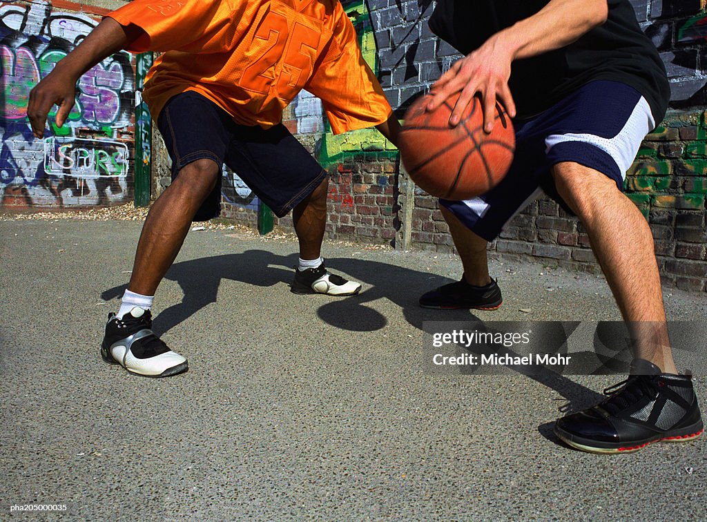Man dribbling basketball against opponent next to graffiti wall