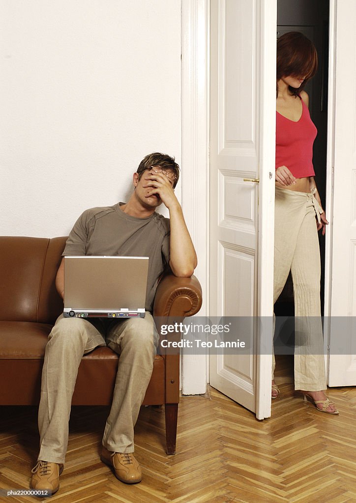Young man using laptop, woman walking into room.