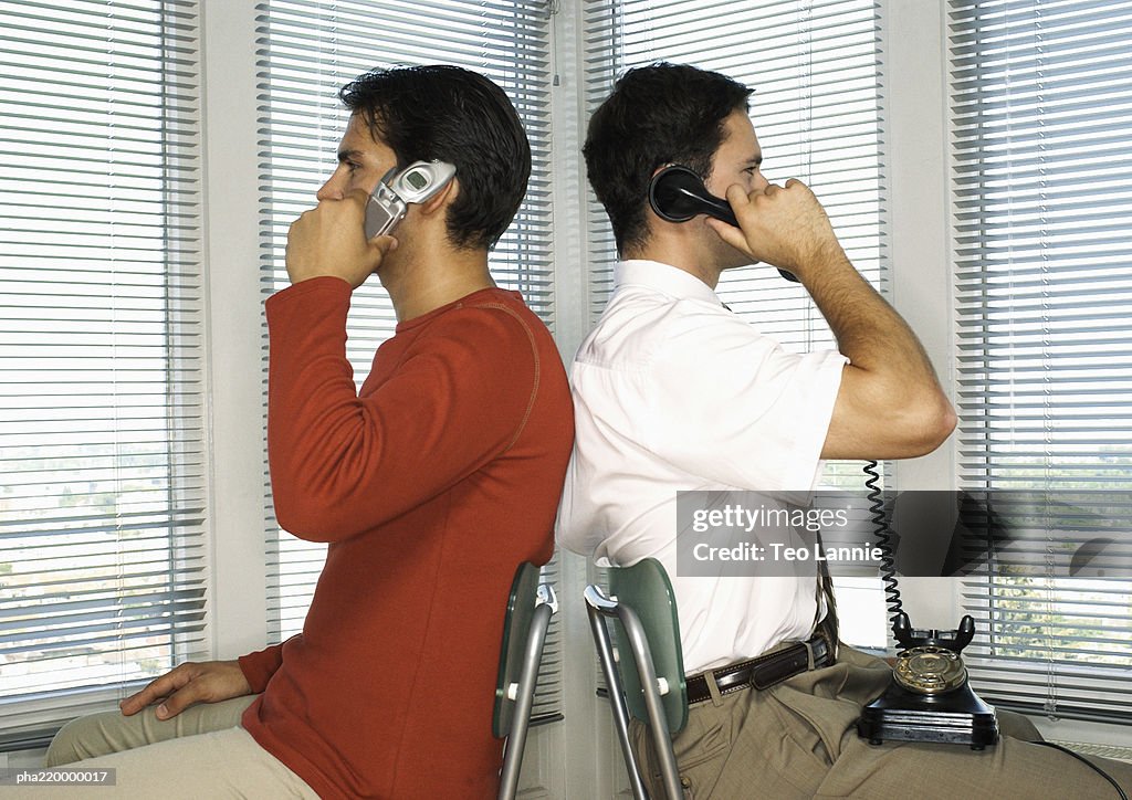 Two men sitting back to back, one with cell phone, other with telephone.