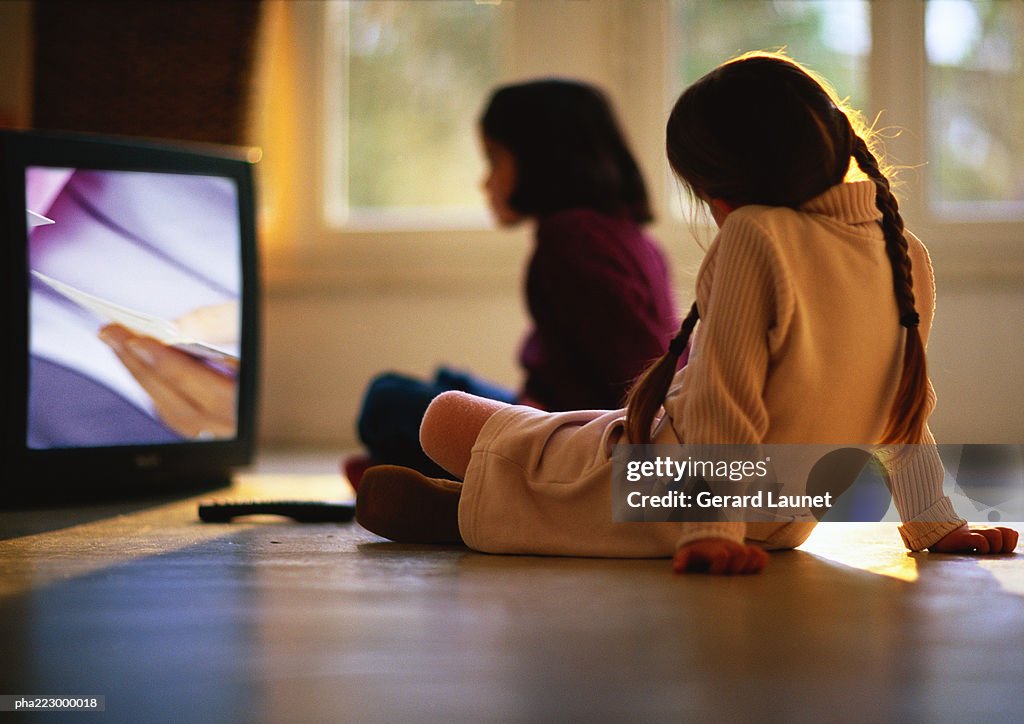 Young girls sitting on wood floor watching TV, girl in background blurred.