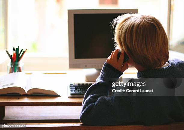 young boy sitting in front of computer talking on phone, view from behind. - reference book stock pictures, royalty-free photos & images