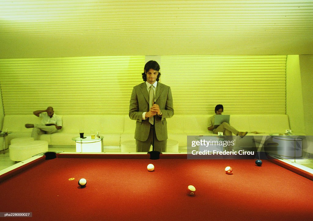 Young man with cuestick, standing at pool table, pool table in foreground, men sitting in background