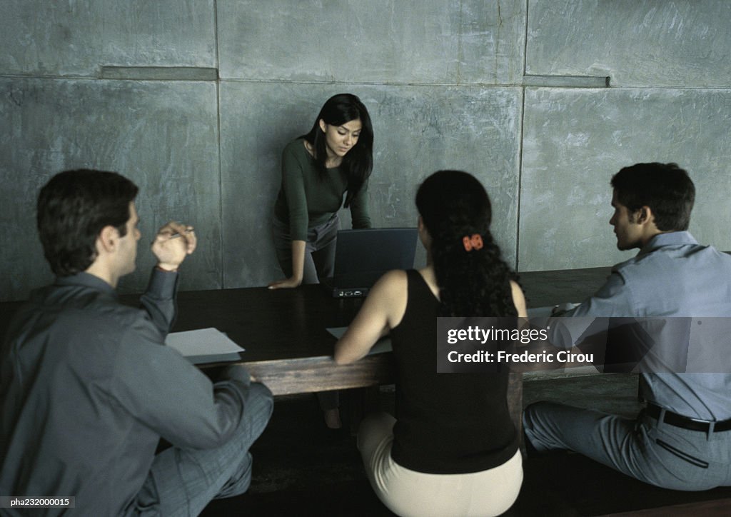 Woman standing at table looking at computer, three people sitting at table.