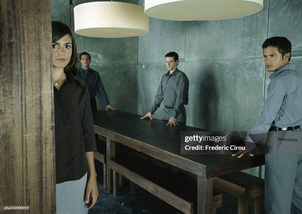 Men standing at table looking at woman standing looking at camera.