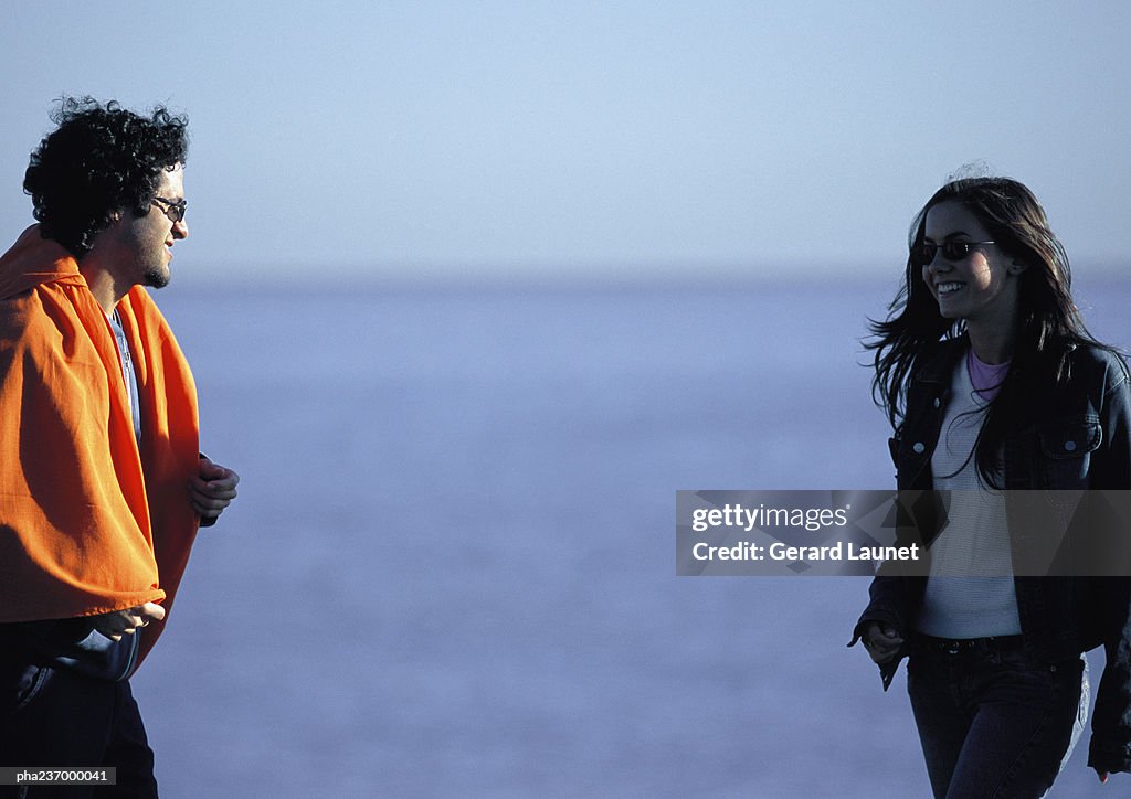 Man wearing orange blanket looking at woman near water.
