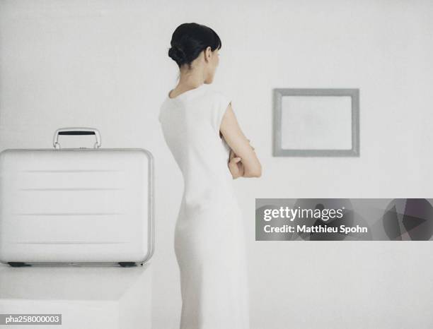 woman standing next to table with metallic briefcase looking toward frame on wall - blank frame stockfoto's en -beelden