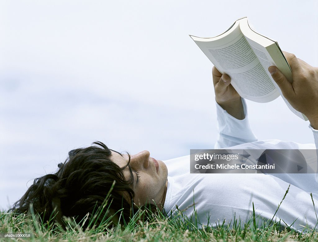 Young man lying on back in grass reading book, head and shoulders