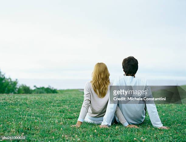young man and young woman sitting side by side on grass, propped up with hands, rear view - propped stock pictures, royalty-free photos & images