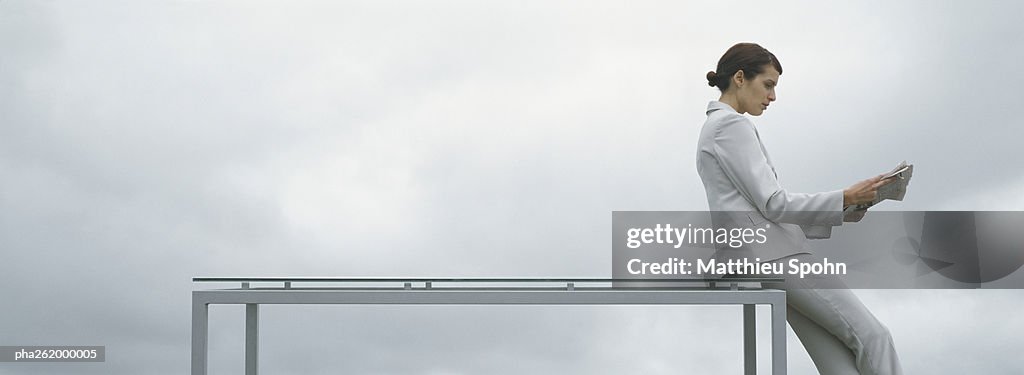 Woman leaning against edge of table, holding newspaper, side view, in front of overcast sky