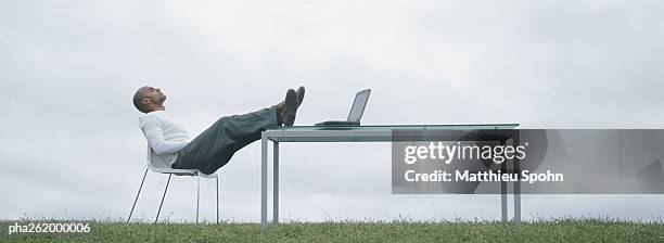 man sitting in chair outdoors with feet on table and laptop on table, in front of overcast sky - legs on the table foto e immagini stock