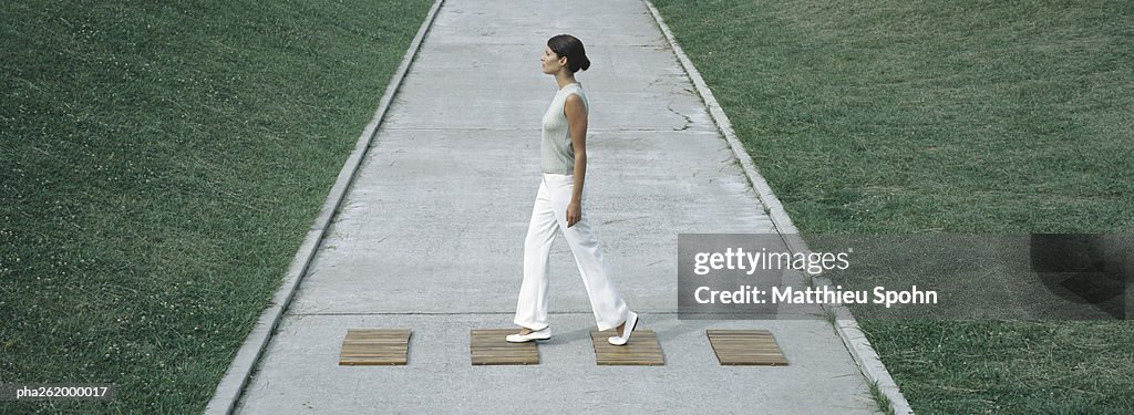 Woman stepping on wood squares across concrete path, full length