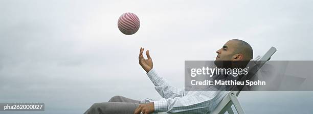 man sitting outdoors in lounge chair throwing ball into air, side view - ball chair stock pictures, royalty-free photos & images