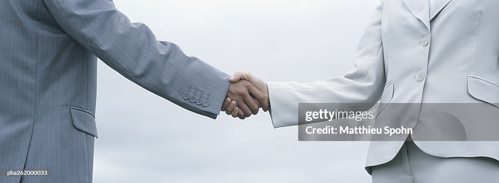 Woman and man in suits shaking hands, mid-section with sky in background