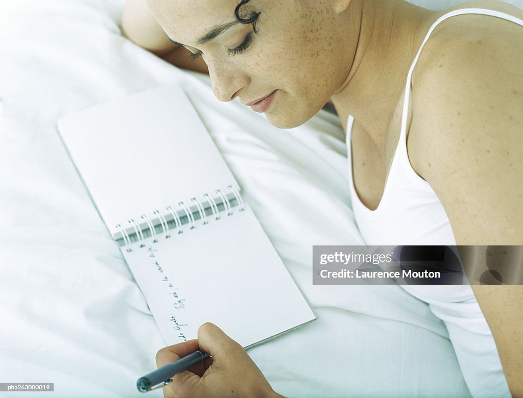 Woman lying on side on bed writing in notebook with pen