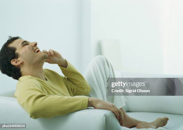 man sitting barefoot on couch talking on phone, laughing with head back - armlehne stock-fotos und bilder