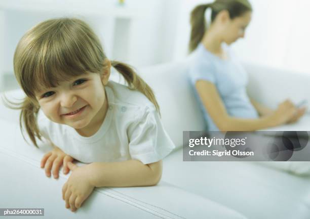 little girl leaning on sofa arm making faces at camera, young woman looking down in background - armlehne stock-fotos und bilder