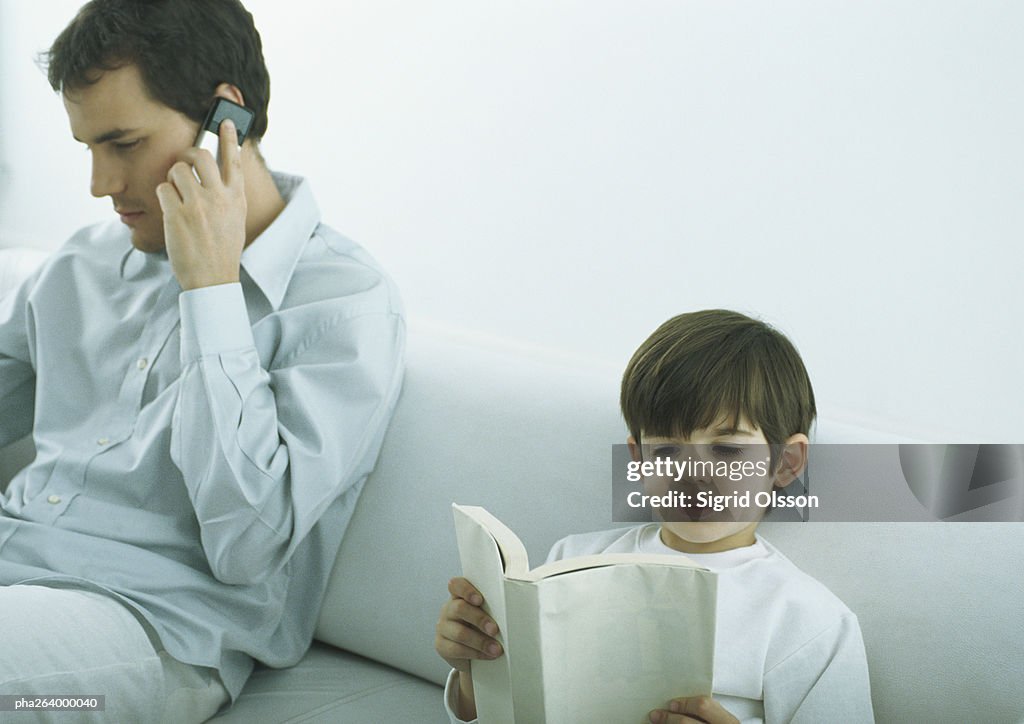 Man sitting on sofa talking on cell phone, boy sitting next to him reading book