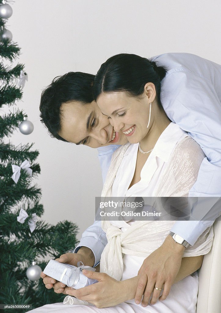 Woman holding gift, man bending over her shoulder, Christmas tree in background