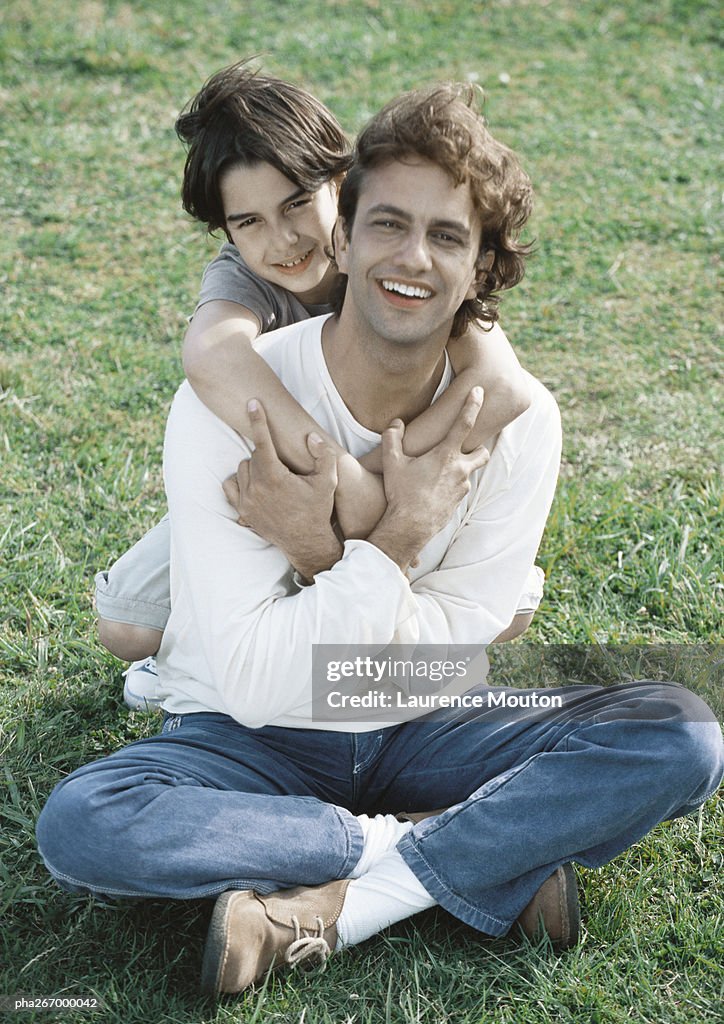 Man sitting on ground, son behind him with arms around man, both looking at camera