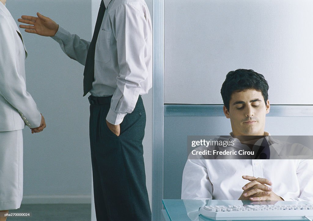 Businessman sitting back at desk with eyes closed, behind him businesspeople standing together, mid section