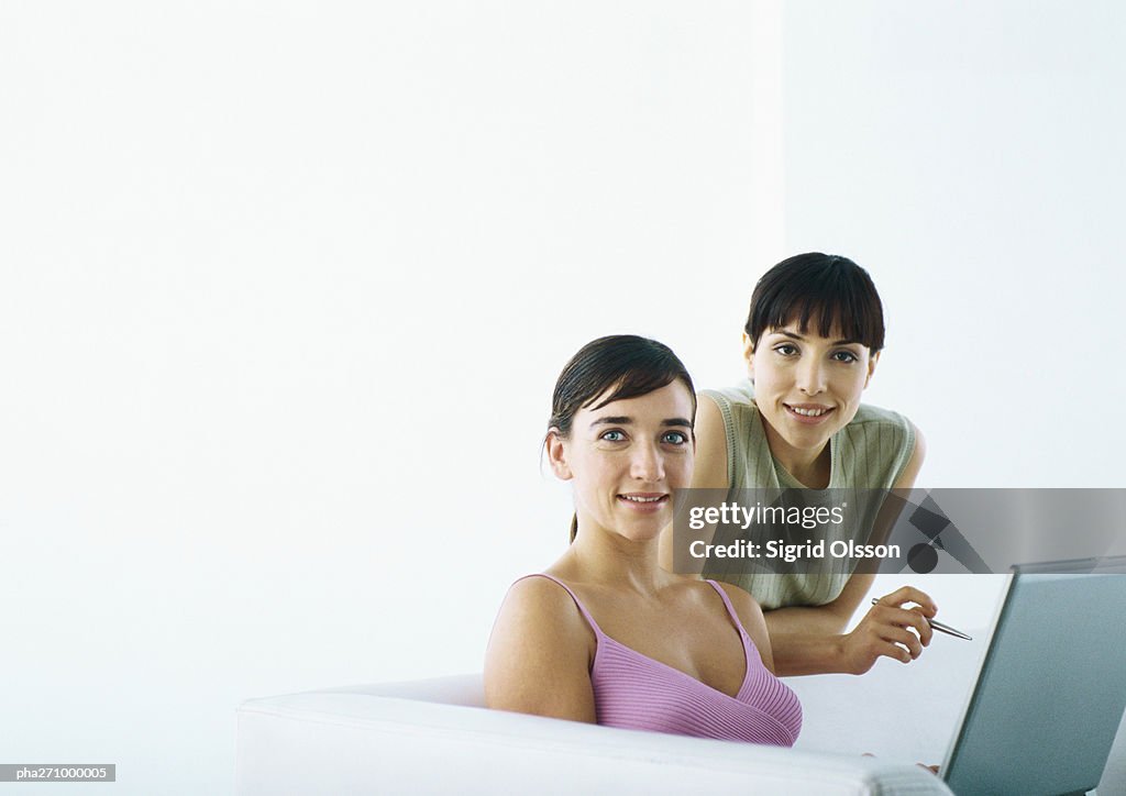 Woman sitting on sofa with laptop, second woman behind leaning on sofa