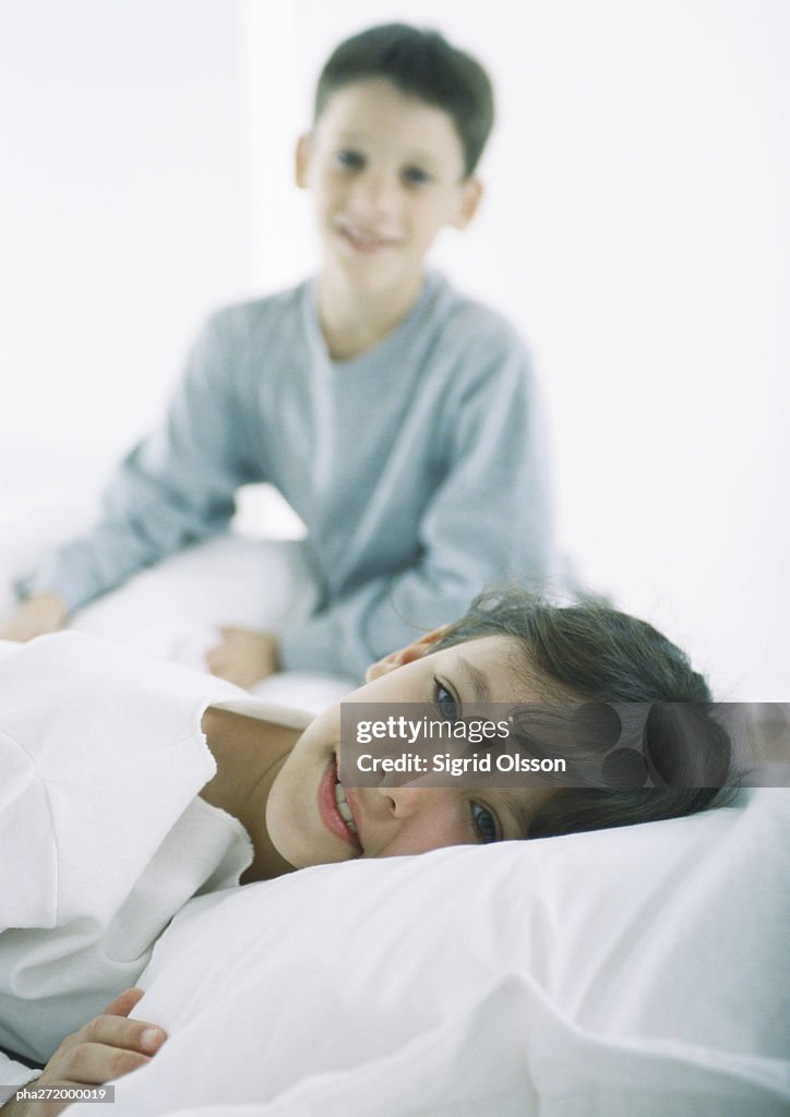 Girl lying in bed looking at camera, boy in background