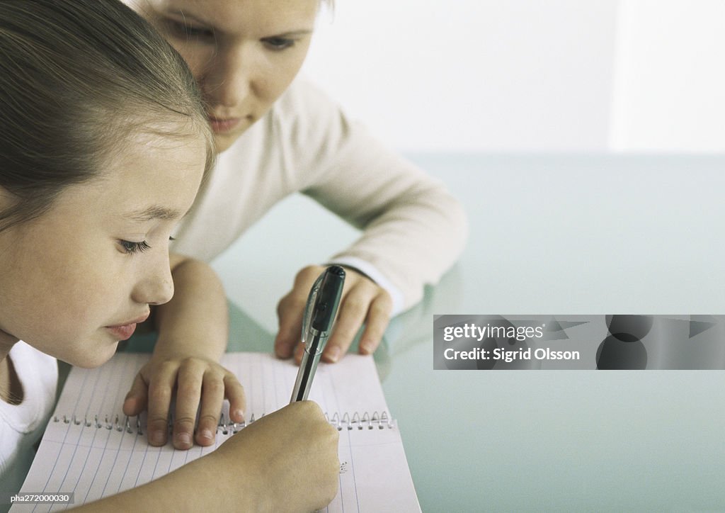 Girl sitting at table writing in notebook, woman at her side