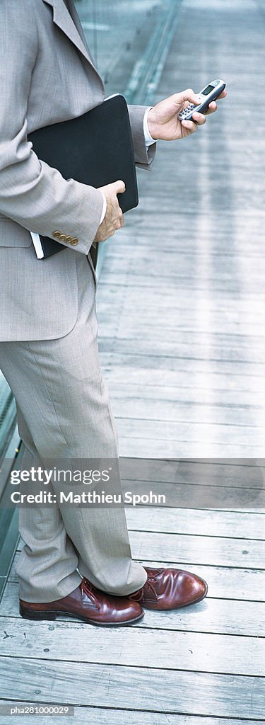 Businessman standing, holding file and dialing cell phone, chest down, panoramic