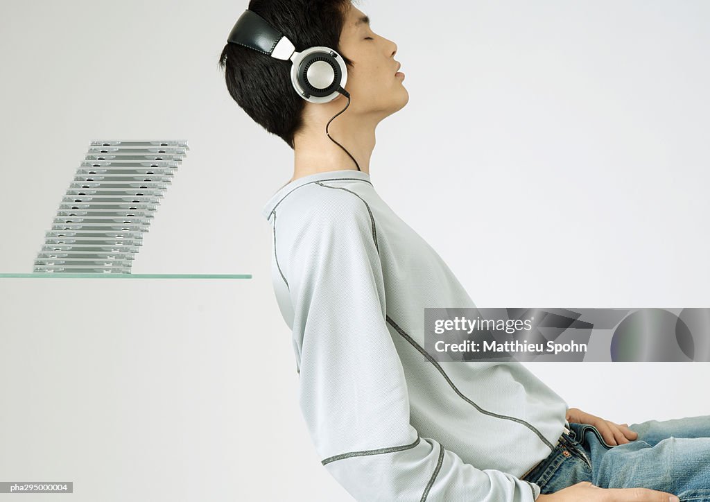 Young man listening to headphones, sitting in front of stack of cds