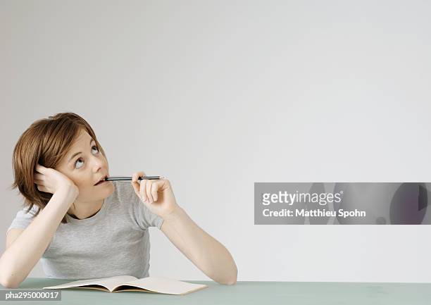 young woman sitting with open notebook, holding pencil to mouth - kin in de hand stockfoto's en -beelden