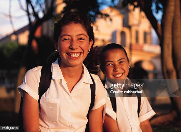 two young latin american schoolgirls with white uniforms and backpacks are smiling into the camera in costa rica - costa stock-fotos und bilder