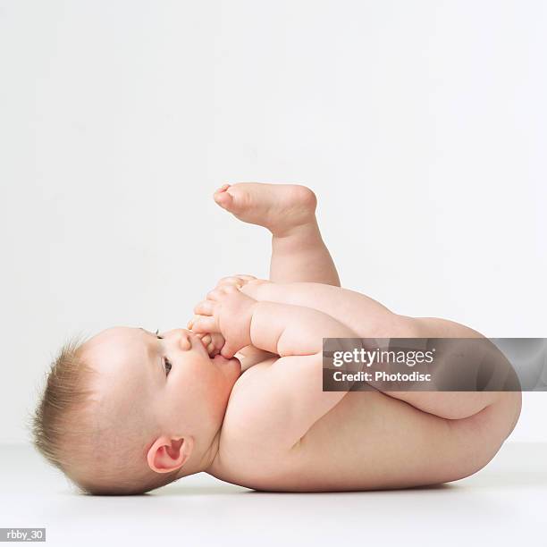 a caucasian baby lies naked on its back and puts its foot in its mouth - feet sucking stock pictures, royalty-free photos & images