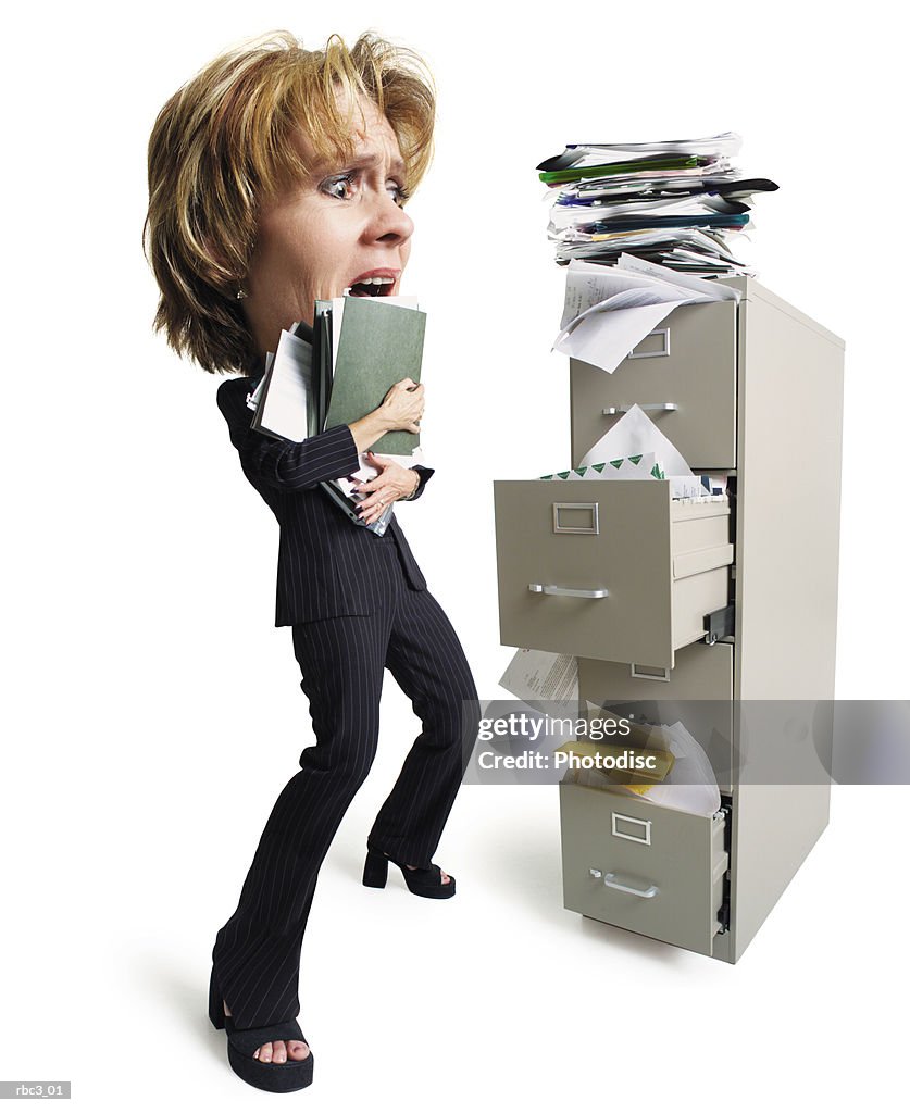 A caucasian woman holding a stack of files stands in front of an overfilled file cabinet looking overwhelmed at all of the papers