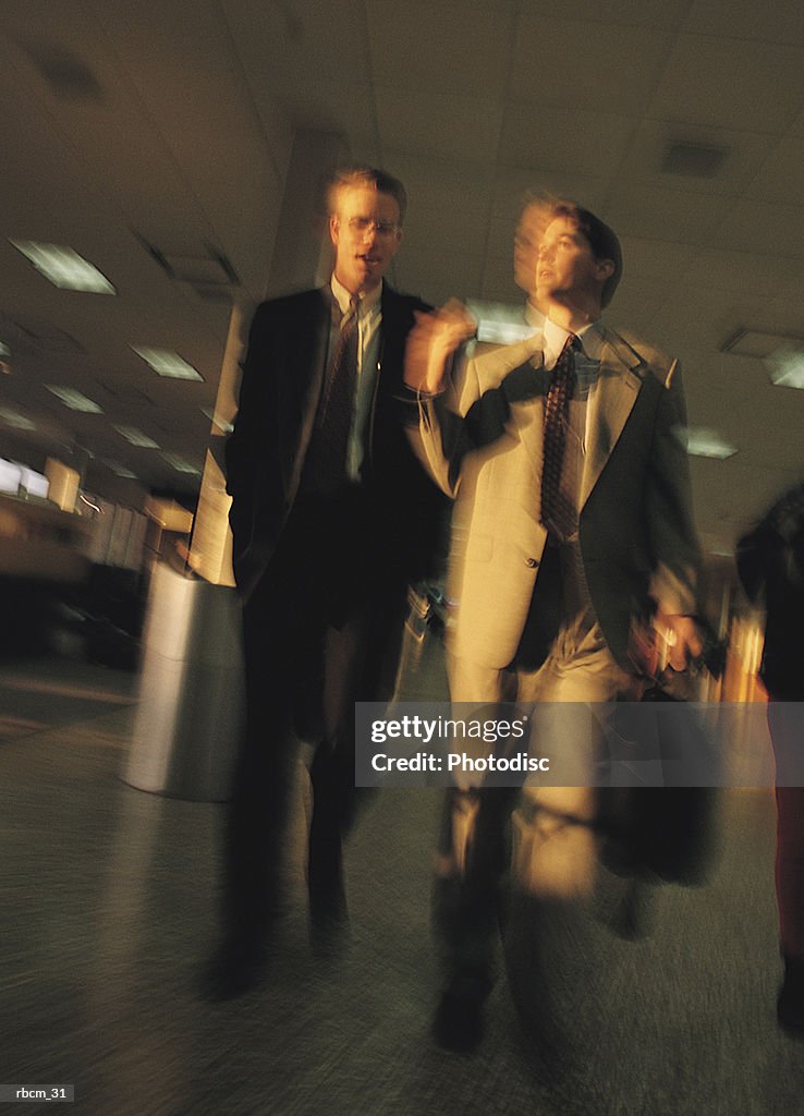 TWO BUSINESSMEN DRESSED IN SUITS TALK AS THEY WALK THROUGH AN AIRPORT CARRYING THEIR BRIEFCASES