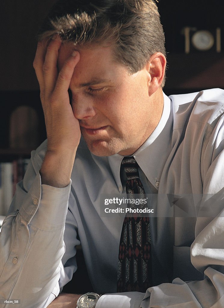 A BUSINESSMAN IN A BLUE SHIRT AND TIE COVERS HIS FACE IN FRUSTRATION