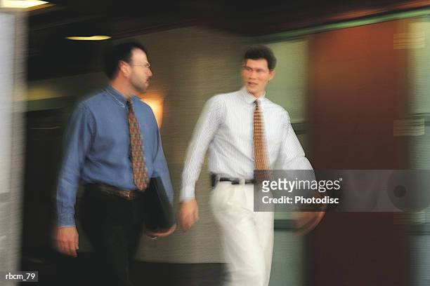 a businessman dressed in a blue shirt and black pants talks to another dressed in a white shirt and pants as they walk down an office hall - aother stock pictures, royalty-free photos & images