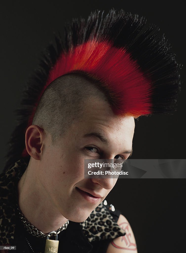A male caucasian teenager with a red and black mohawk is dressed in punk clothes and smirking
