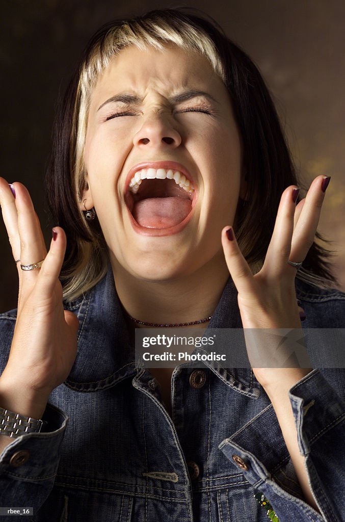 A caucasian young woman with brown and blond streaked hair wearing a jean jacket is screaming with eyes closed and hands raised near her face