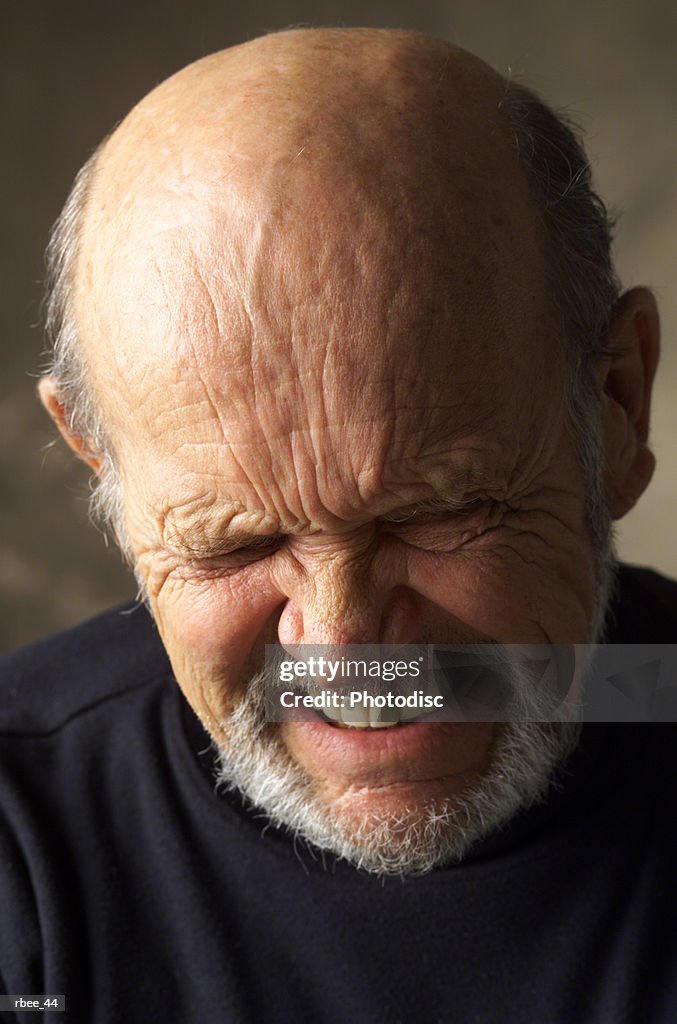 A wrinkle faced balding elderly man wearing a dark shirt scrunches up his face while closing his eyes and grimacing