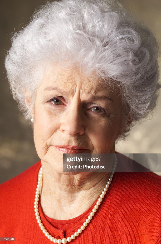An elderly caucasian woman with curly white hair is wearing a red shirt and has an expression of sadness