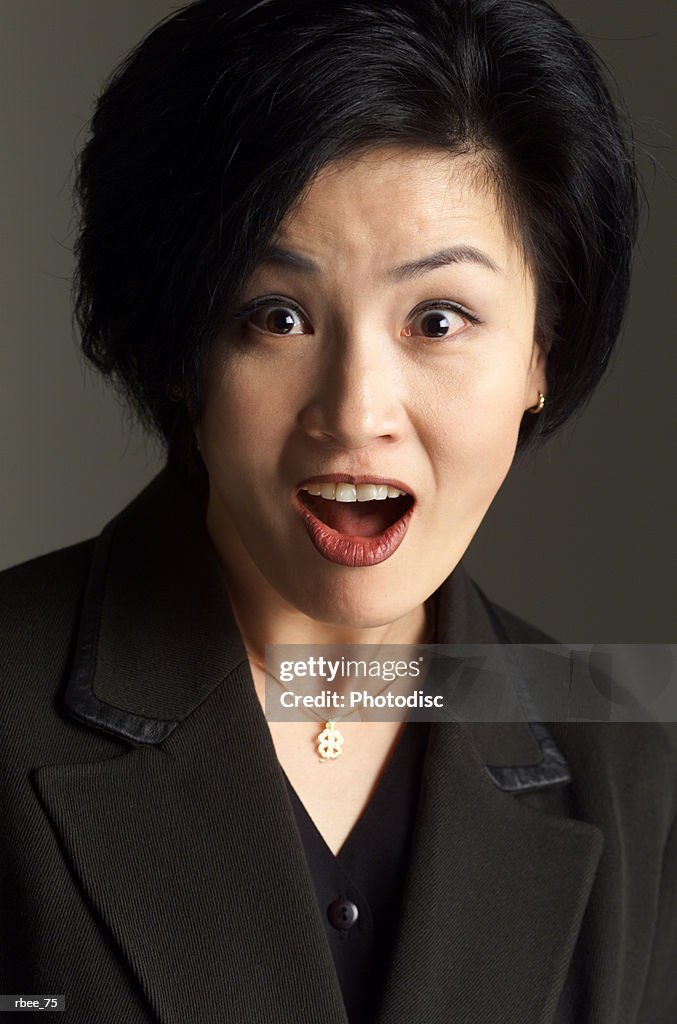 An asian woman wearing a dark suit has short hair and is staring open mouthed with an expression of shock