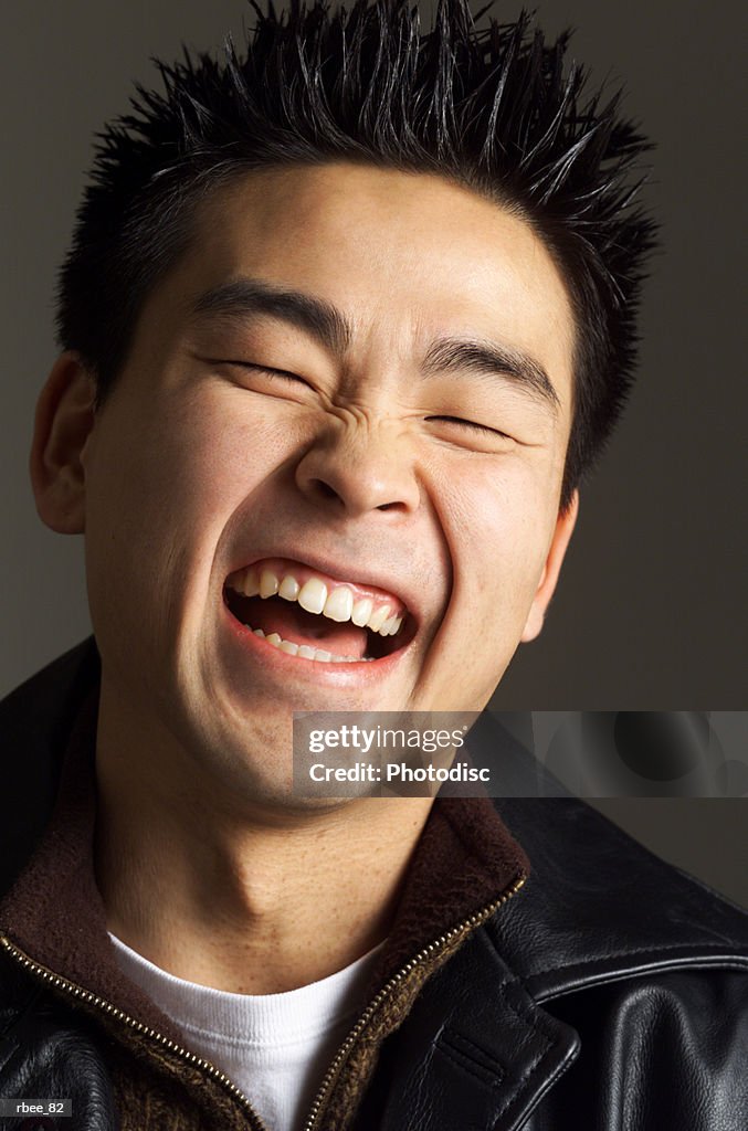 A young asian man with spiked hair and a leather jacket is laughing open mouthed with his eyes shut