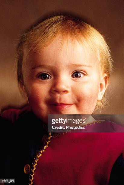 baby girl with short strawberry blonde hair and big blue eyes wearing a red and black shirt smiles softly to someone with an innocent and happy look on her face - happy stockfoto's en -beelden