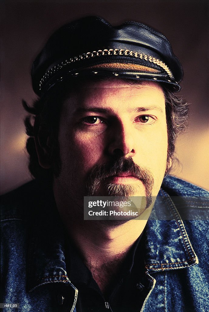 A man with a dark mustache and dark hair wearing a leather hat with a chain on it and a denim jacket stares at the camera with little emotion and appears to be a tough biker or motorcyclist