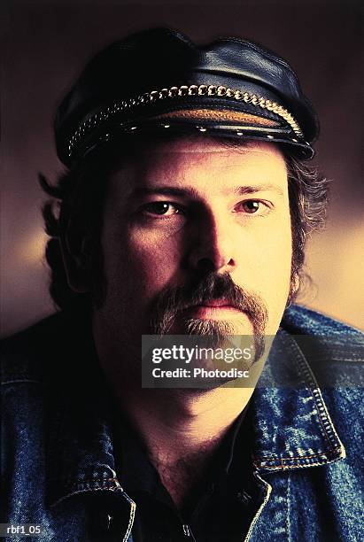a man with a dark mustache and dark hair wearing a leather hat with a chain on it and a denim jacket stares at the camera with little emotion and appears to be a tough biker or motorcyclist - handlebar moustache stock pictures, royalty-free photos & images